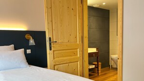 Une chambre avec une porte en bois, un lit avec une literie blanche, une table de chevet avec une lampe, et une vue sur la salle de bain.