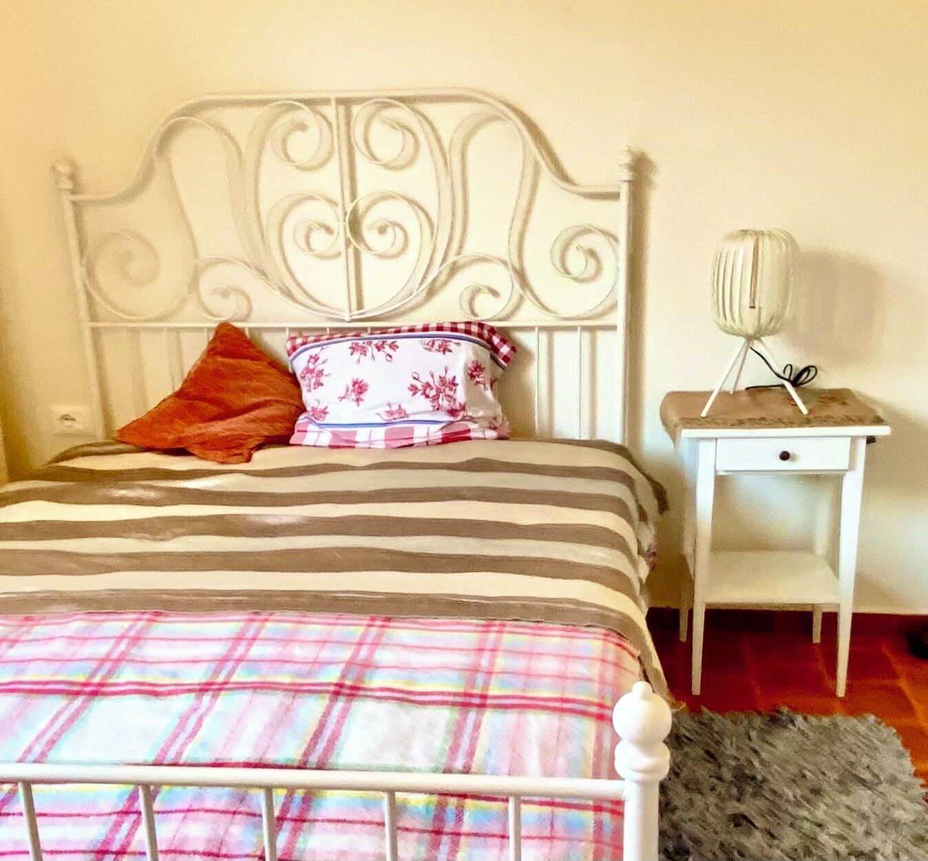 Un lit avec une tête de lit en métal, une couverture à rayures et un coussin à motifs floraux. Il y a une petite table de chevet avec une lampe et un tiroir.