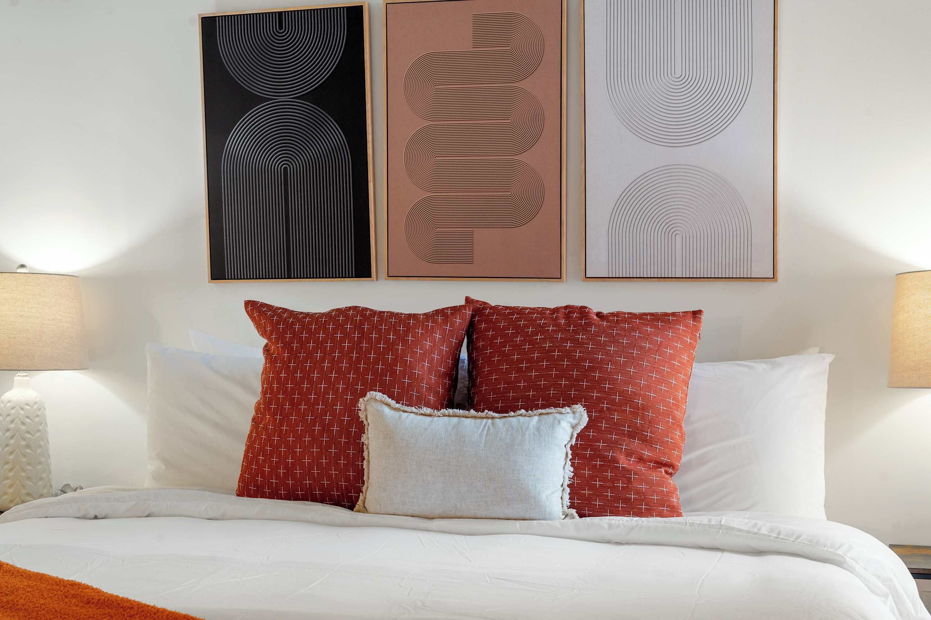 A neatly made bed with red and white patterned pillows, a white pillow, and a white duvet. There are three framed artworks above the bed, each with a unique abstract design.