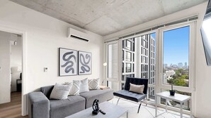 A modern living room with a grey sofa, a white coffee table, and a black chair. There are abstract wall art pieces and a large window with a city view. - New 4 King Bed / Family Ready Apartment (Chicago)