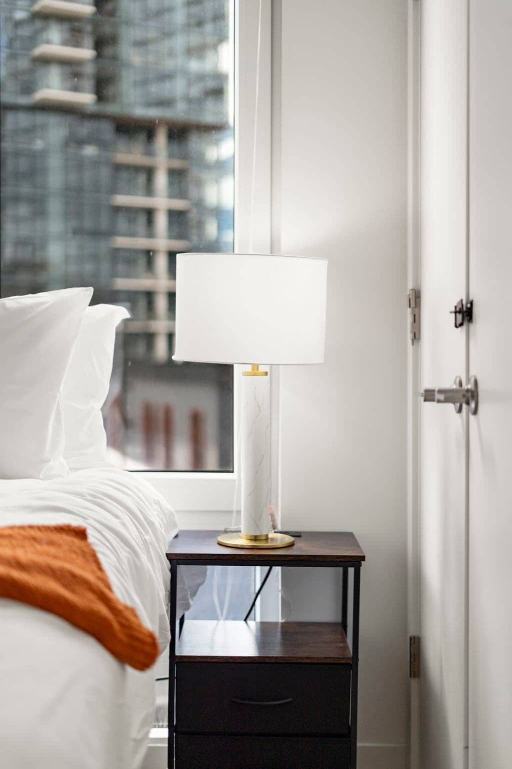 A neatly made bed with white linens and a brown throw, a bedside table with a white lamp, and a view of a cityscape through a window.