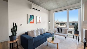 A modern living room with a blue sofa, a round coffee table, and a large window offering a city view. - Bright 3BR Stylish Apartment Near North Ave Shops (Chicago)