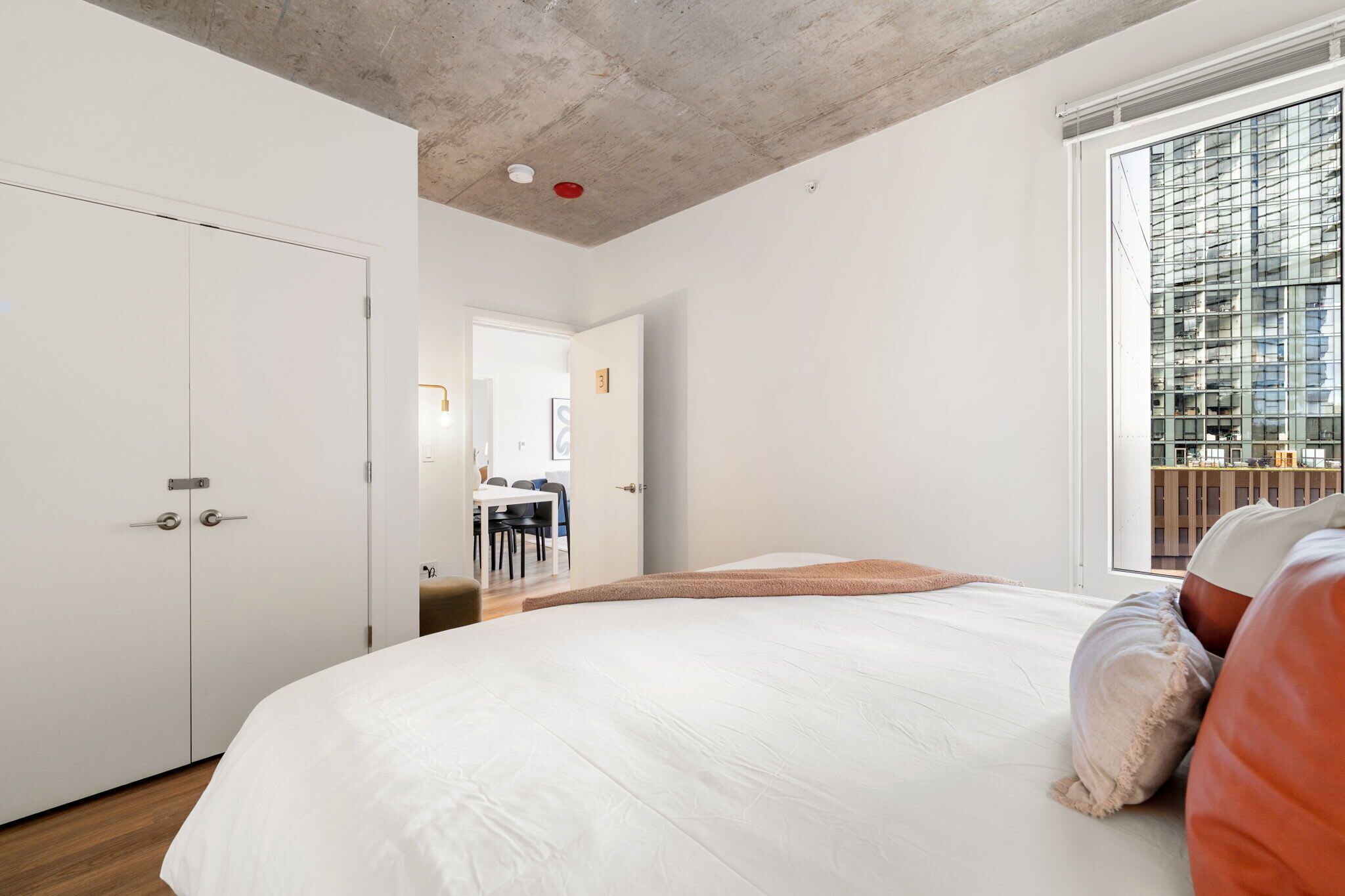 A modern bedroom with a large bed, a wardrobe, and a view of a cityscape through a large window.