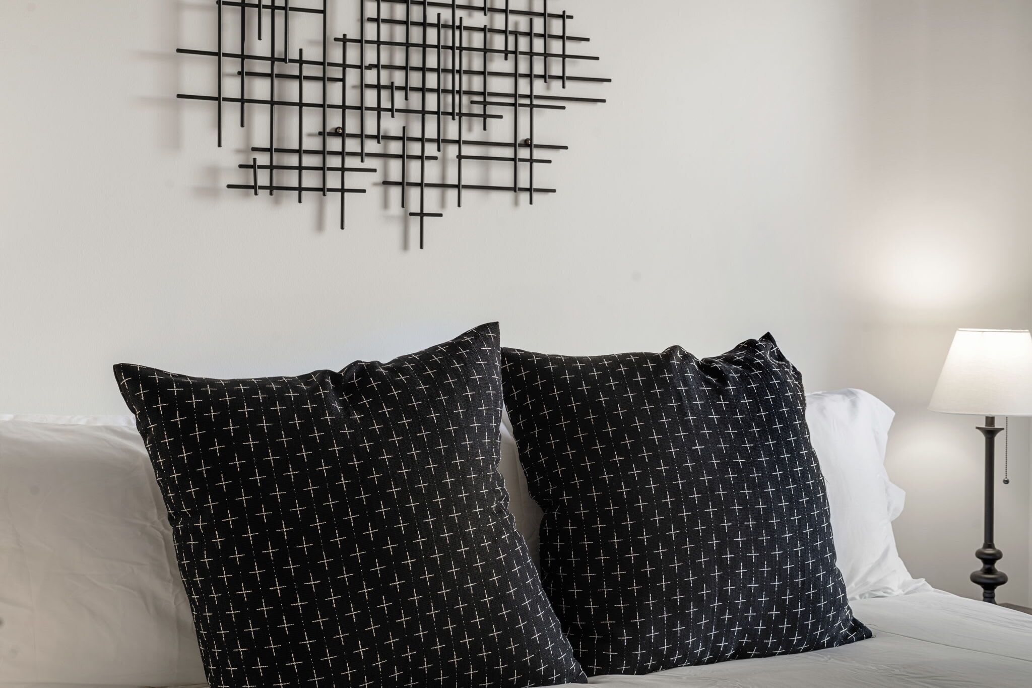 Une chambre d’hôtel avec une œuvre d’art abstraite moderne accrochée au mur, deux coussins à motifs, un lit blanc et une lampe de chevet.