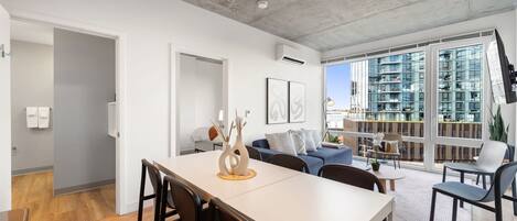 A modern living room with a dining area, large windows, and a view of high-rise buildings.