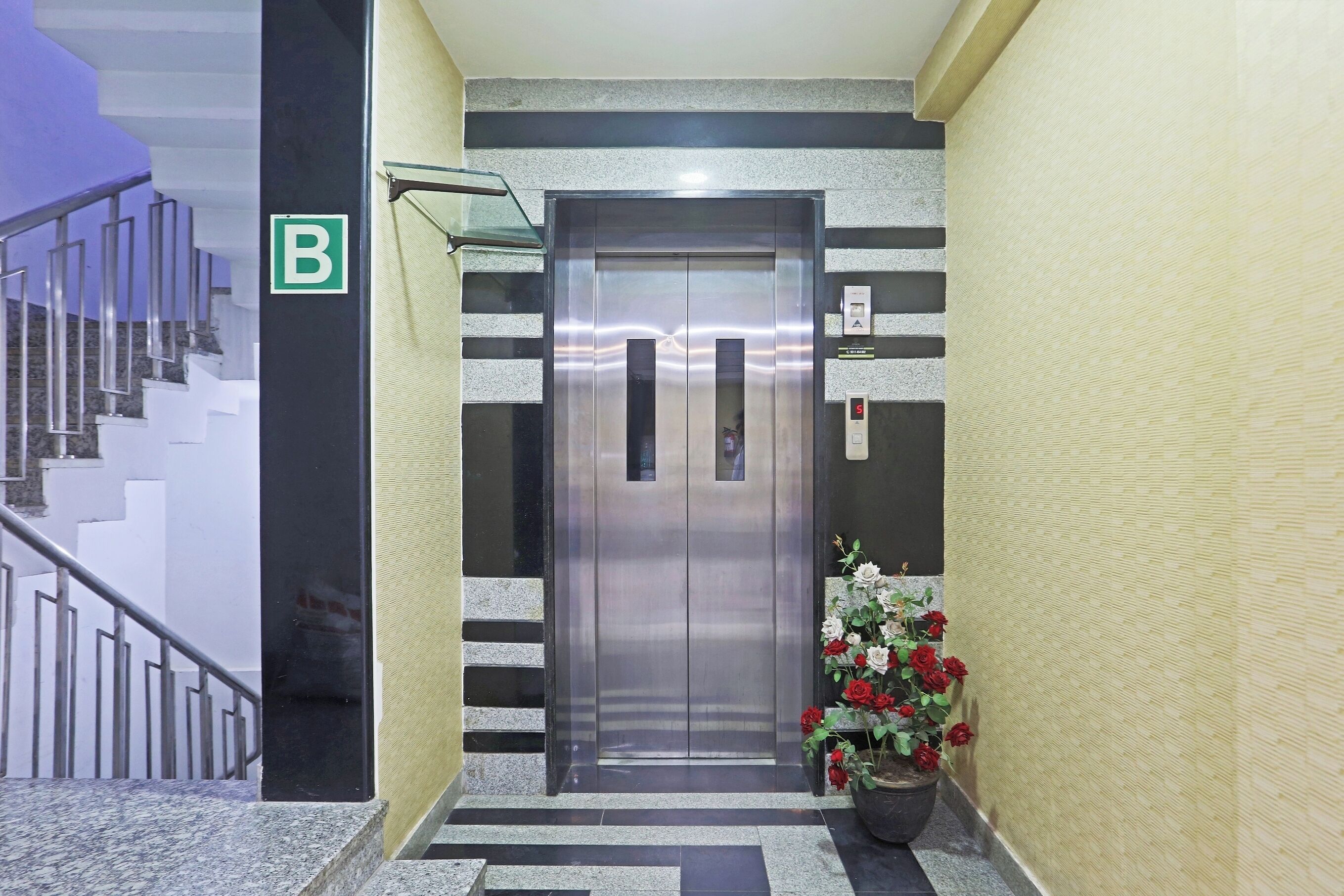 An elevator with a metallic finish, a 'B' sign on the wall, and a potted plant with red and white flowers beside it.