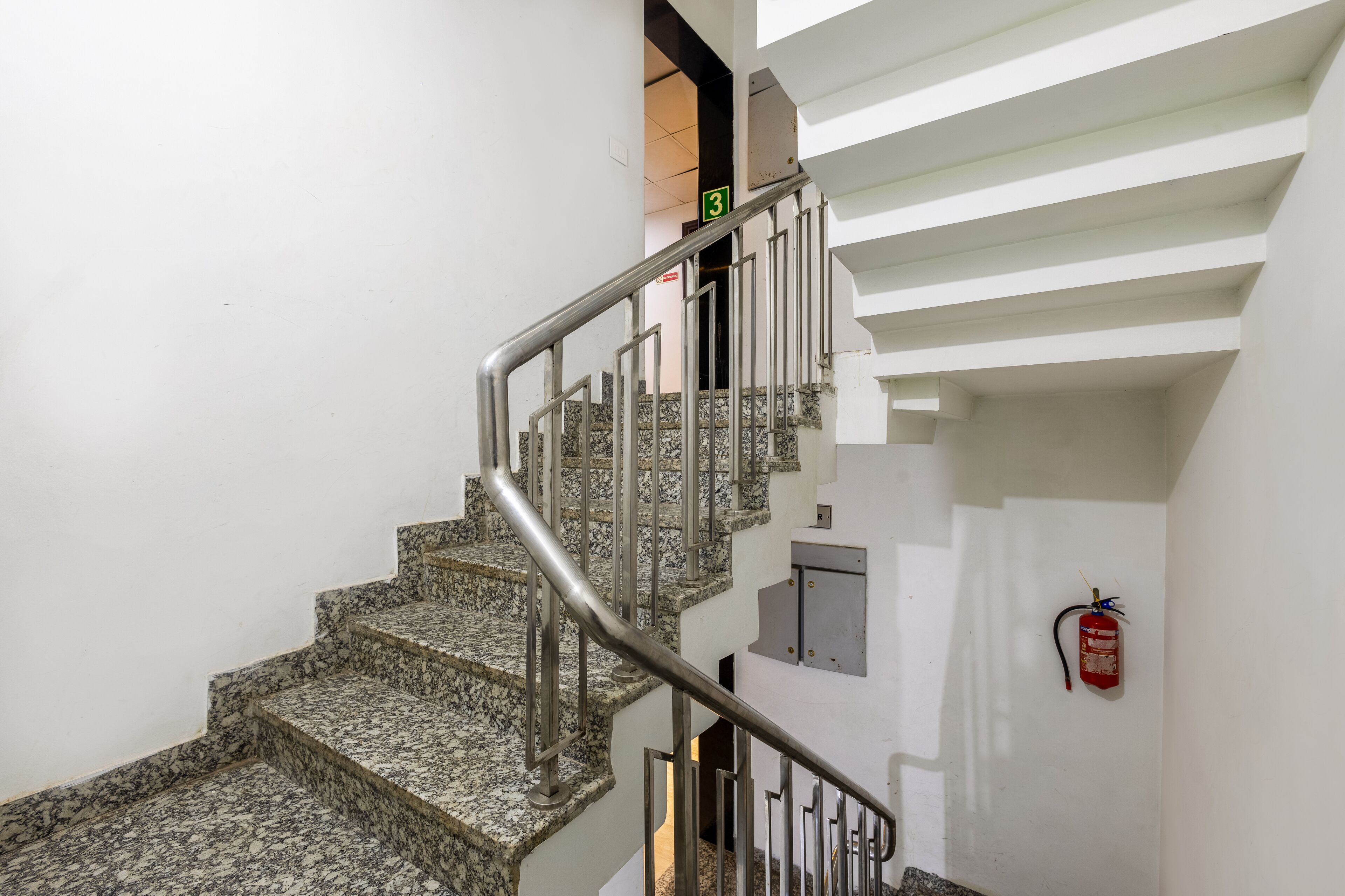 Un escalier avec des marches en granit et des rampes en métal, un extincteur fixé au mur, et un panneau vert indiquant le niveau de l’étage.