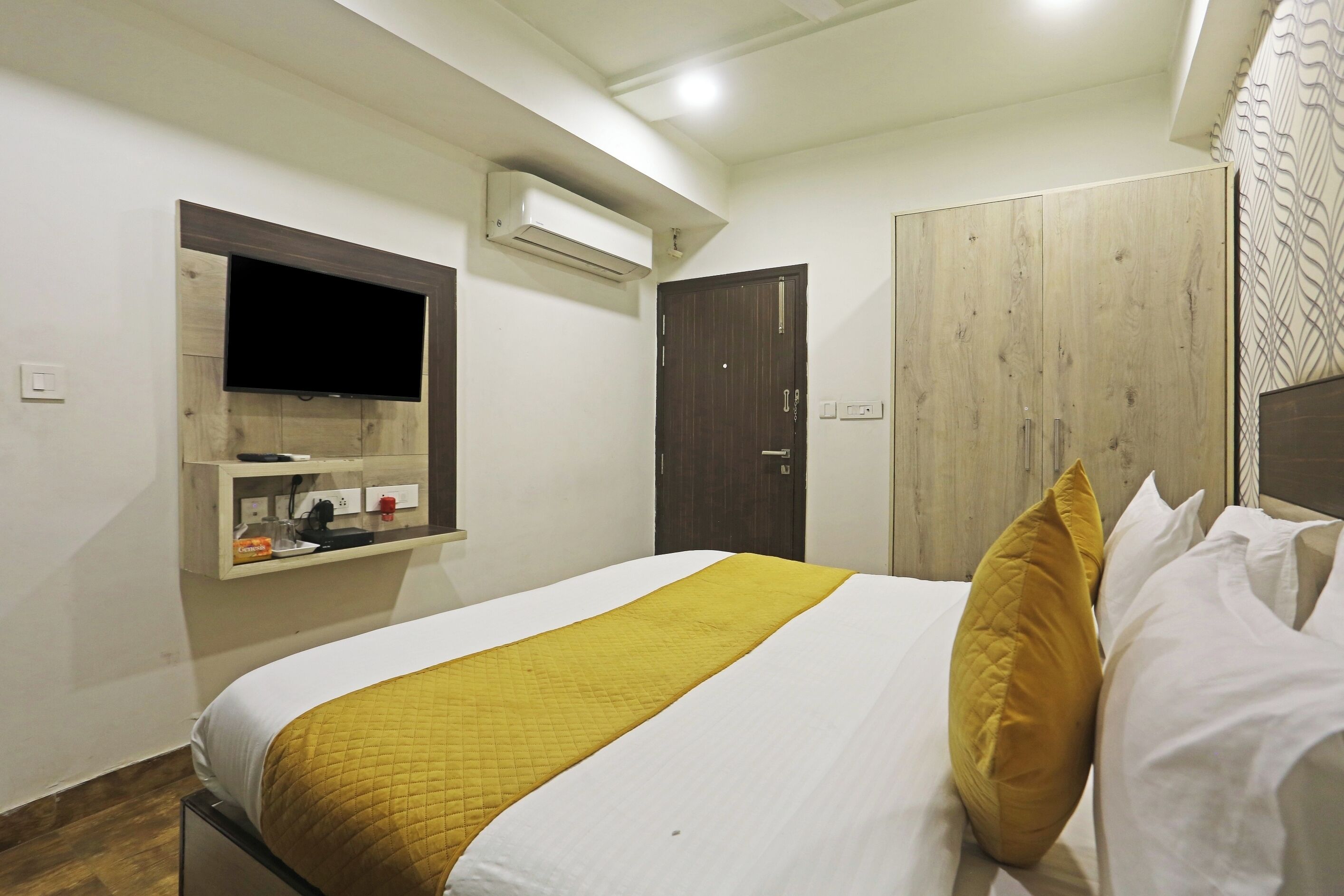 A neatly made bed with a white and yellow bedspread, a wooden wardrobe, a mounted television, and an air conditioning unit on the wall.