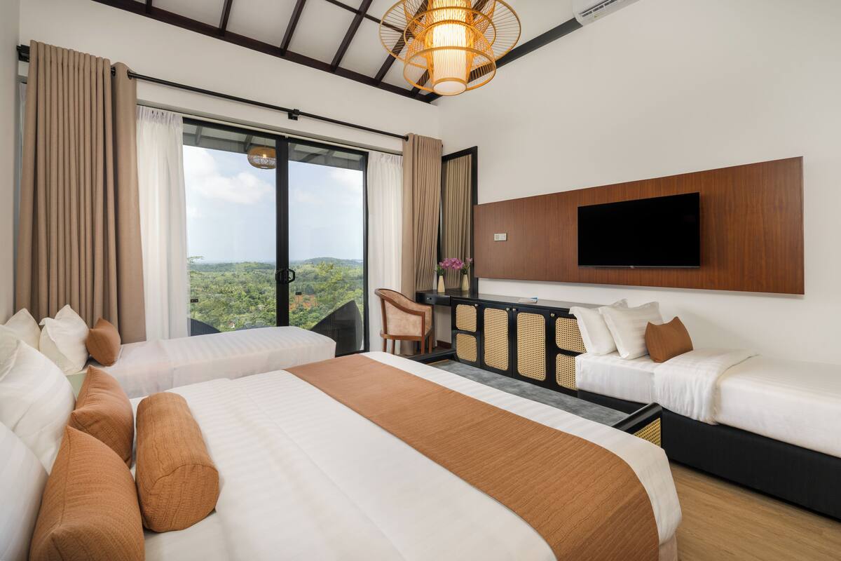 A hotel room with two beds, a TV mounted on the wall, a desk, and a view of the outdoors.