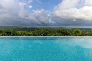 A clear blue infinity pool with a view of a lush green landscape and distant mountains under a cloudy sky.