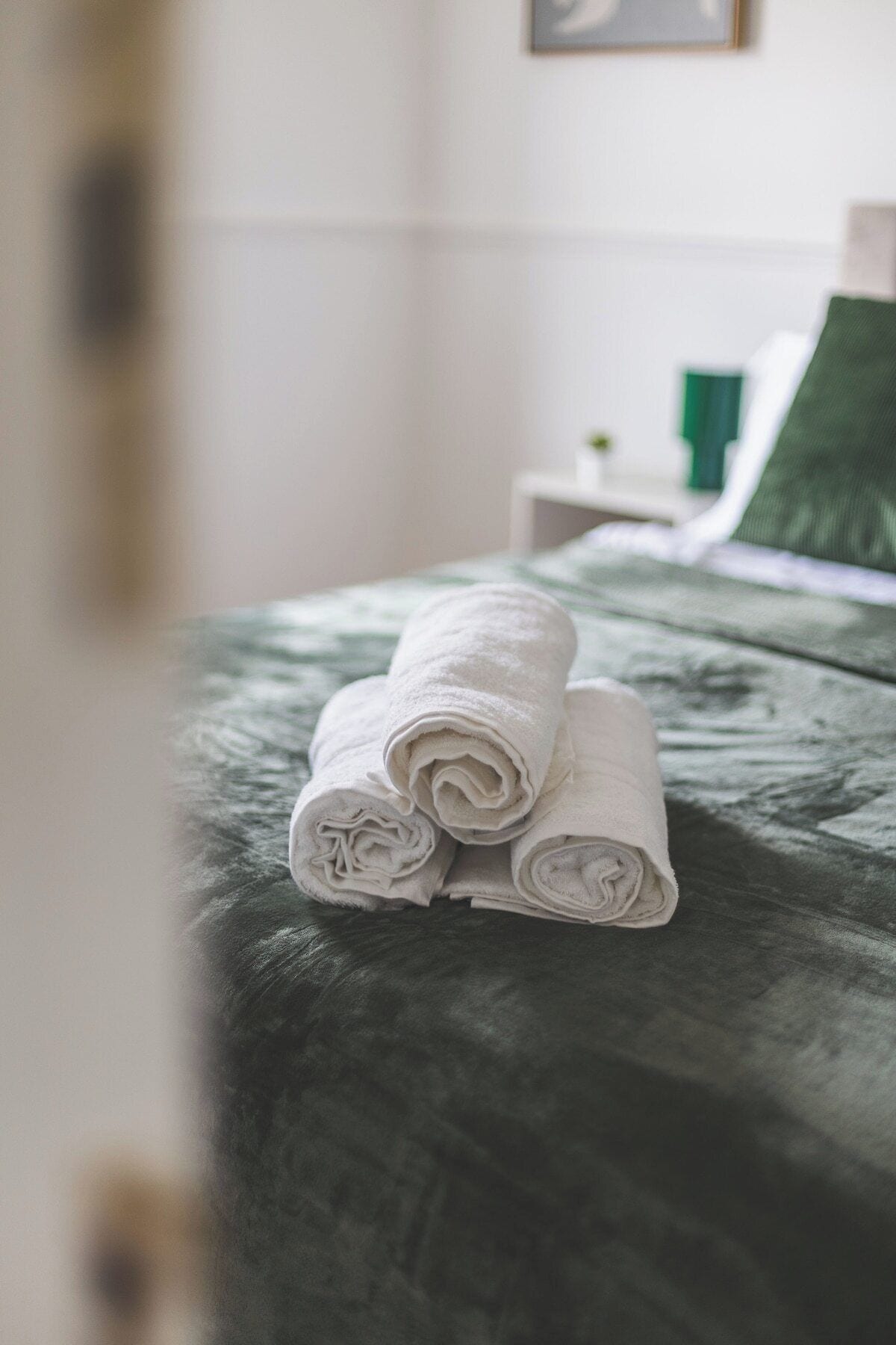 Un lit bien fait, avec des serviettes blanches pliées en rouleaux et une tête de lit verte recouverte de tissu.