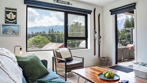 Un salon chaleureux avec une grande fenêtre donnant sur les montagnes, un fauteuil confortable, une table en bois sur laquelle est posée une assiette de poires, et une carte encadrée accrochée au mur.