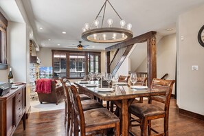 A dining area with a wooden table set for six, surrounded by upholstered chairs. A chandelier hangs from the ceiling, and a stone wall is visible in the background. - Silver Star 104. Pool, Hot Tub, and Plaza Dining Nearby (Park City)