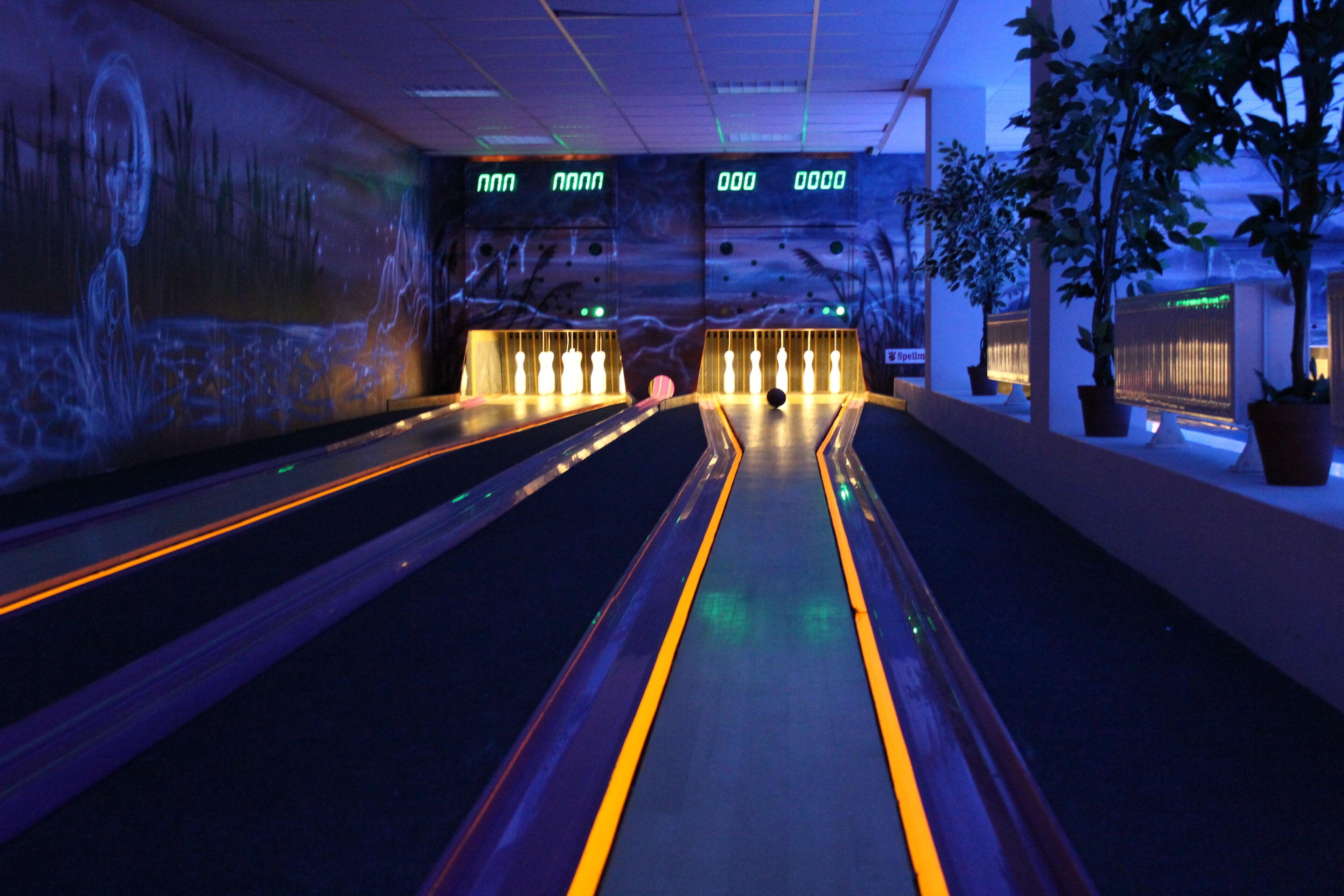 A bowling alley with multiple lanes, illuminated by overhead lights, and a mural on the wall.