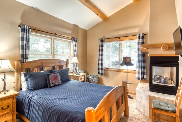 A cozy bedroom with a bed, bedside tables, a lamp, a fireplace, and windows with curtains.