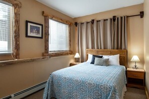 A bedroom with a bed, two bedside tables, a window with blinds, a picture on the wall, and a curtain.