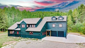 A two-story house with a steep gable roof, multiple windows, and a garage. It is surrounded by a forested area with mountains in the background. - Mountain Meadow Lodge • Dog-Friendly • Hot Tub (Frisco)
