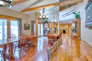 A spacious dining area with wooden chairs and a large table, a kitchen with wooden cabinets, and a view of the outdoors through large windows.
