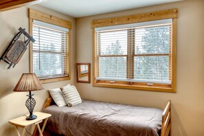 A small bedroom with a single bed, a nightstand, a lamp, and two windows with blinds.