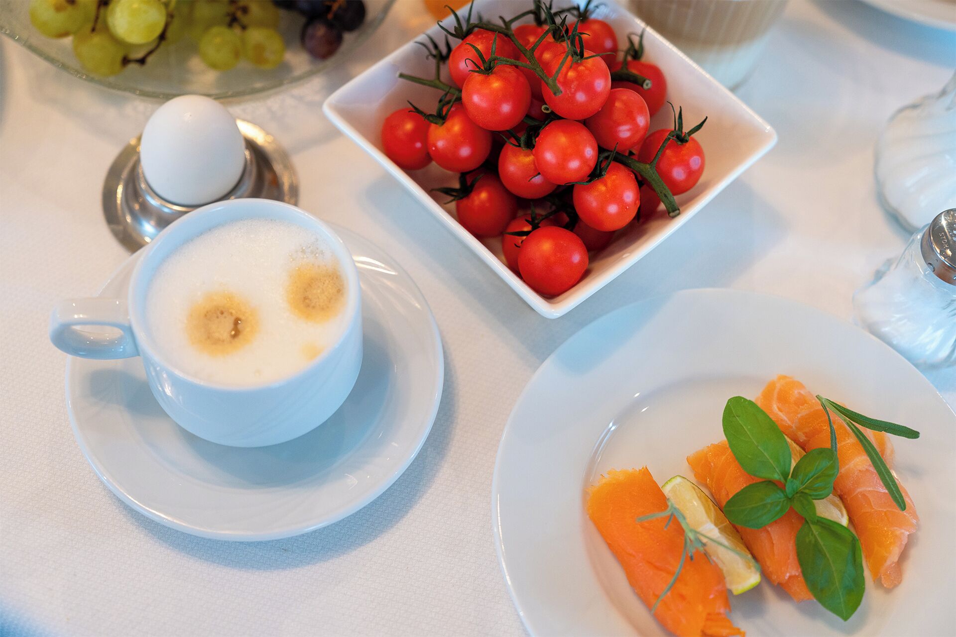 Un desayuno con una taza de café, un bol de tomates cherry y un plato de salmón con limón y hierbas.