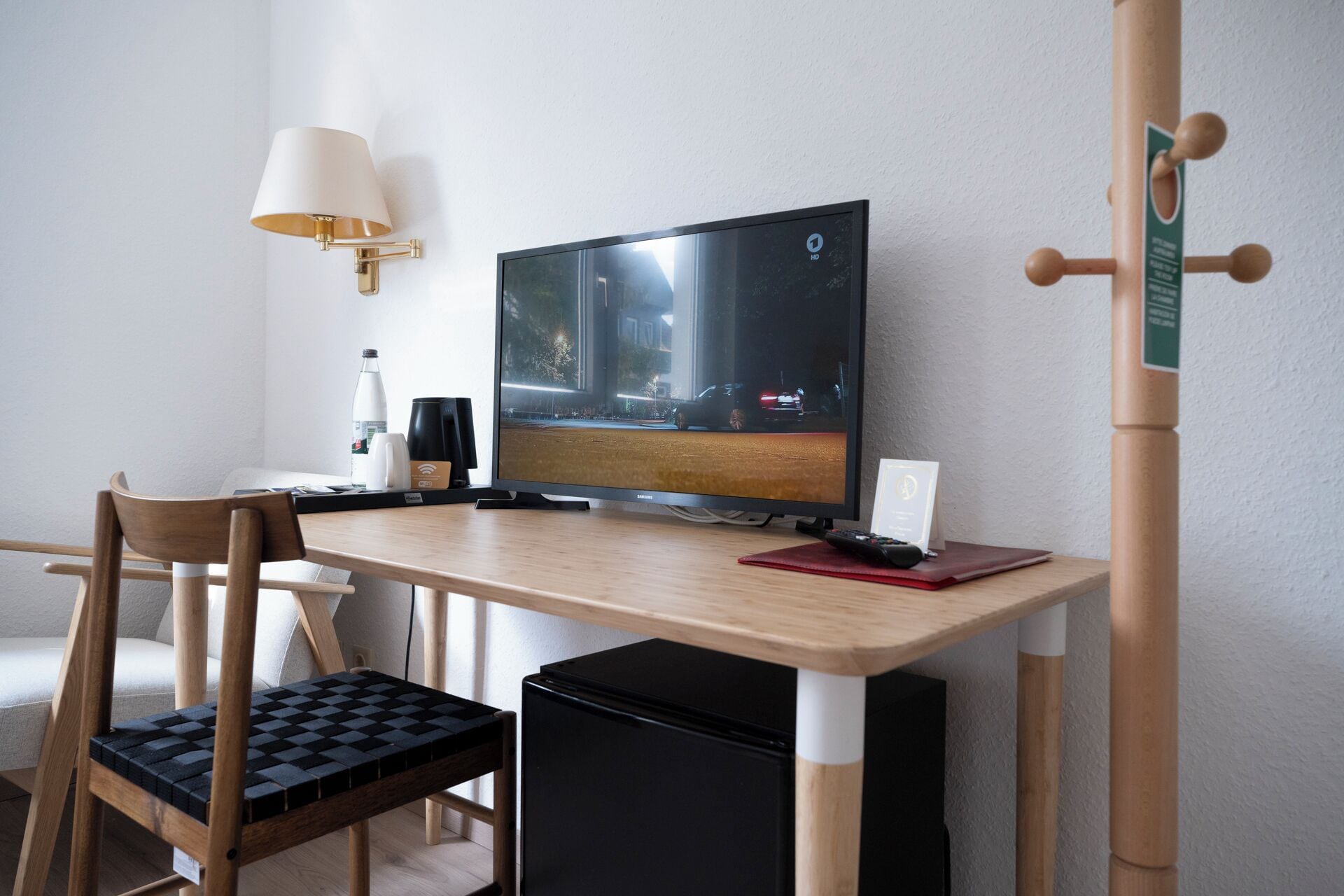 A hotel room with a wooden desk, a flat-screen TV, a chair, a small black cabinet, and a wall-mounted lamp.