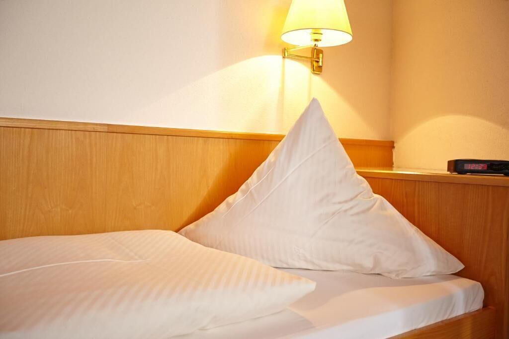 A neatly made bed with white linens and pillows, a bedside table with a lamp, and a digital clock.