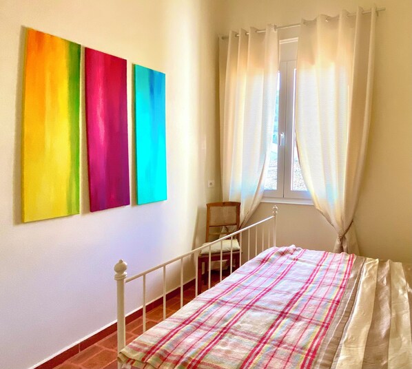 A bedroom with a bed, a window with curtains, and three colorful paintings on the wall.