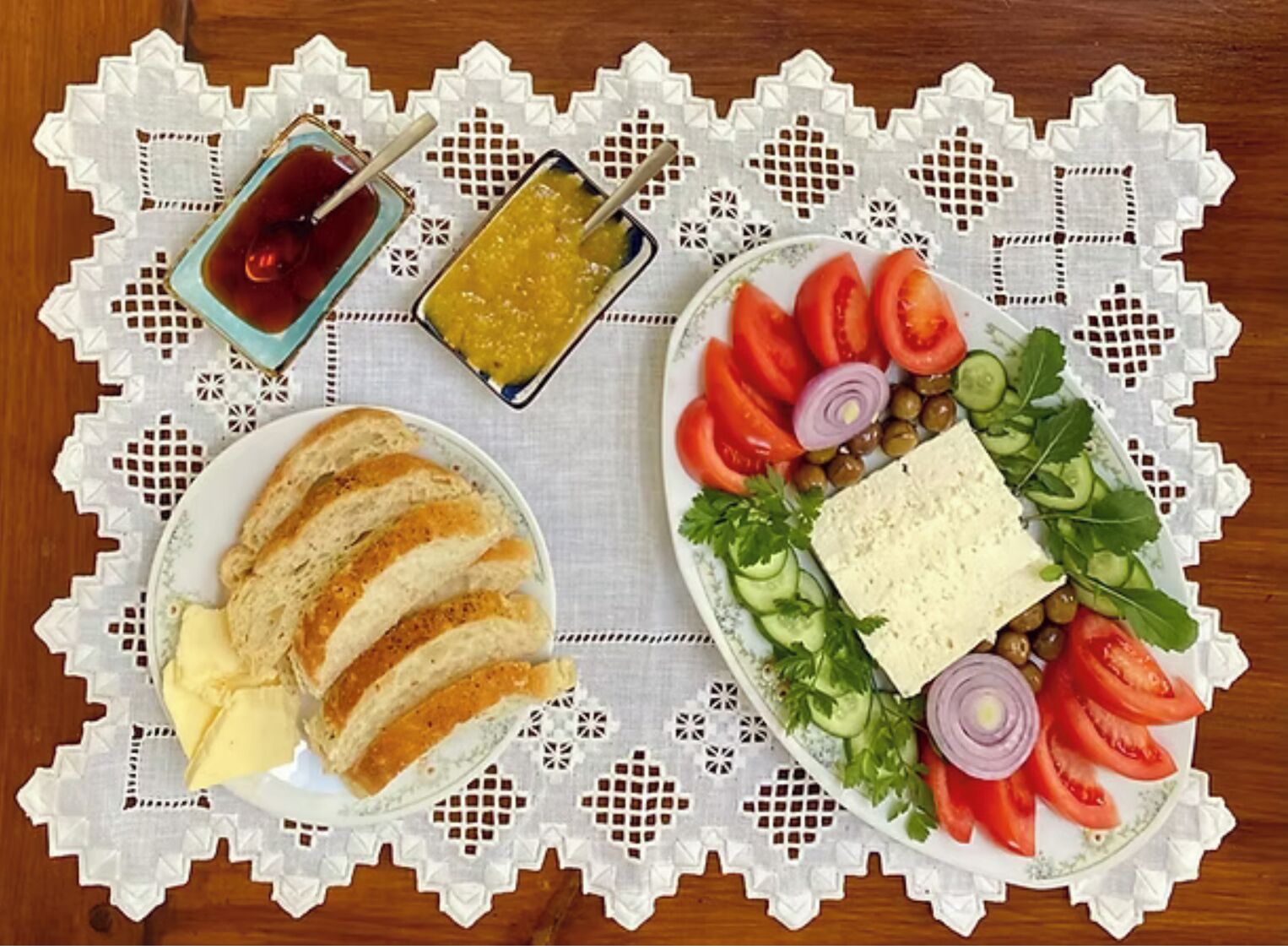 Een tafel met een witte kantkleed, een bord met sneetjes brood en boter, een bord met groenten en kaas, en twee kleine schaaltjes met sauzen.