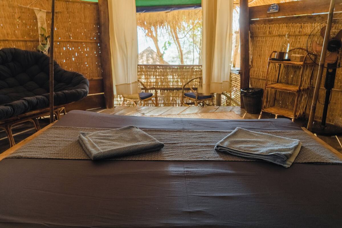 A room with a bed, two chairs, a small table, a fan, and a window with a view of trees.