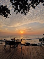 Um deck de madeira com mesa e cadeiras voltado para o oceano ao pôr do sol.
