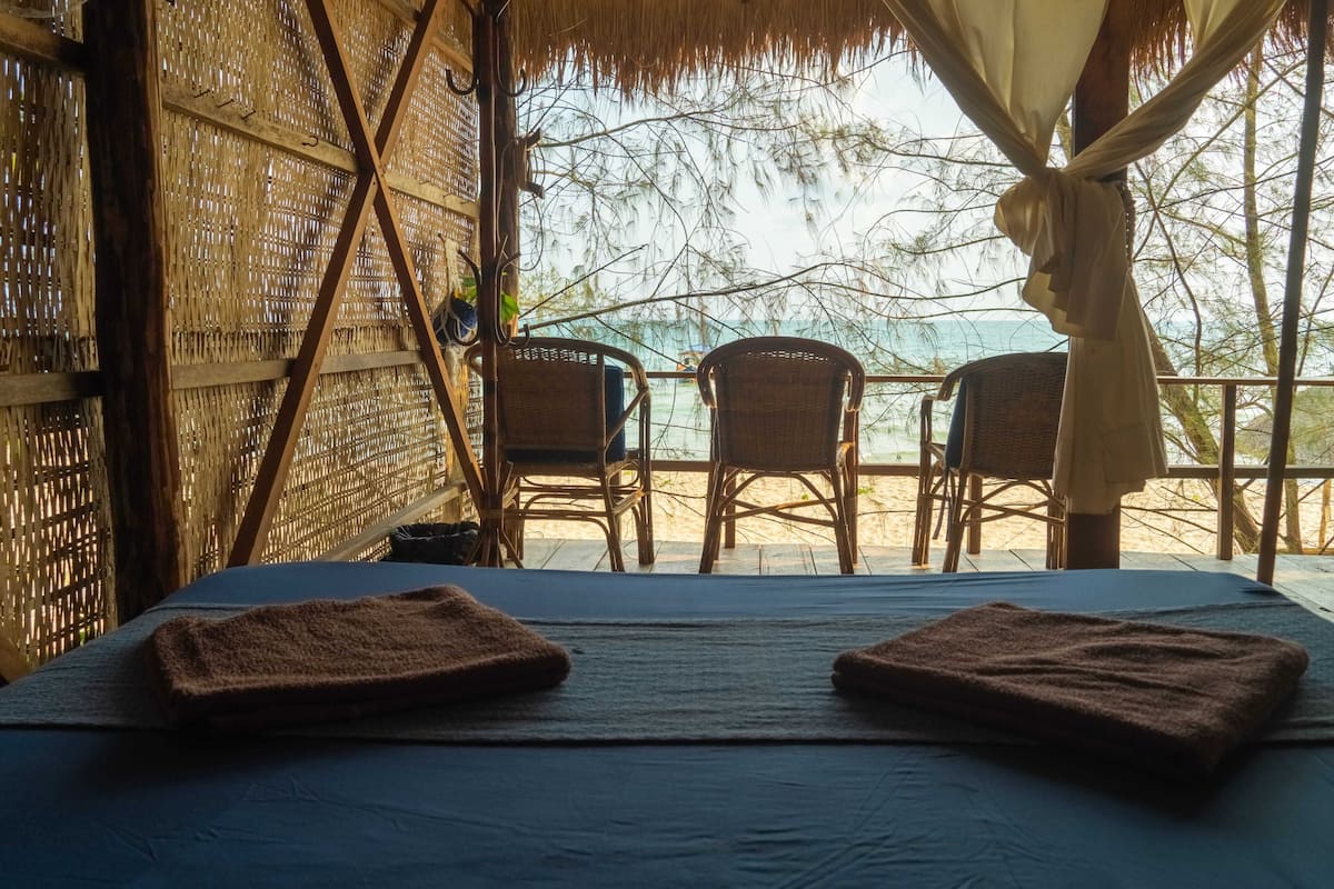A room with a bed, two chairs, a straw wall, and a view of the beach.