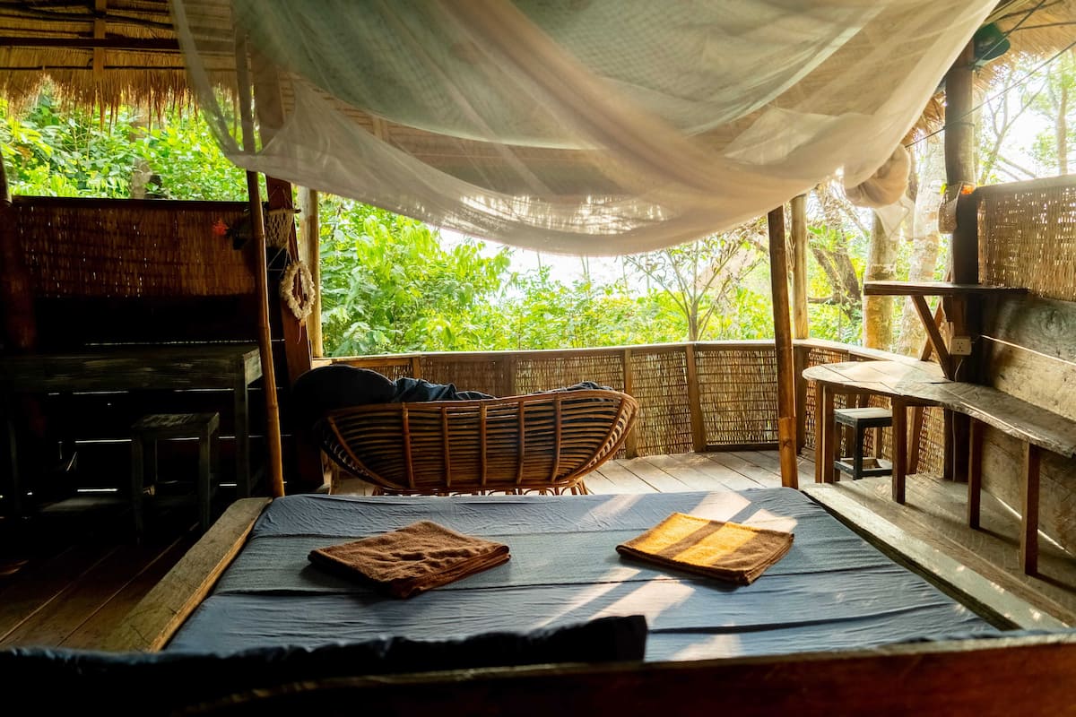 A room with a bed, a wicker chair, a small table, and a view of greenery outside.