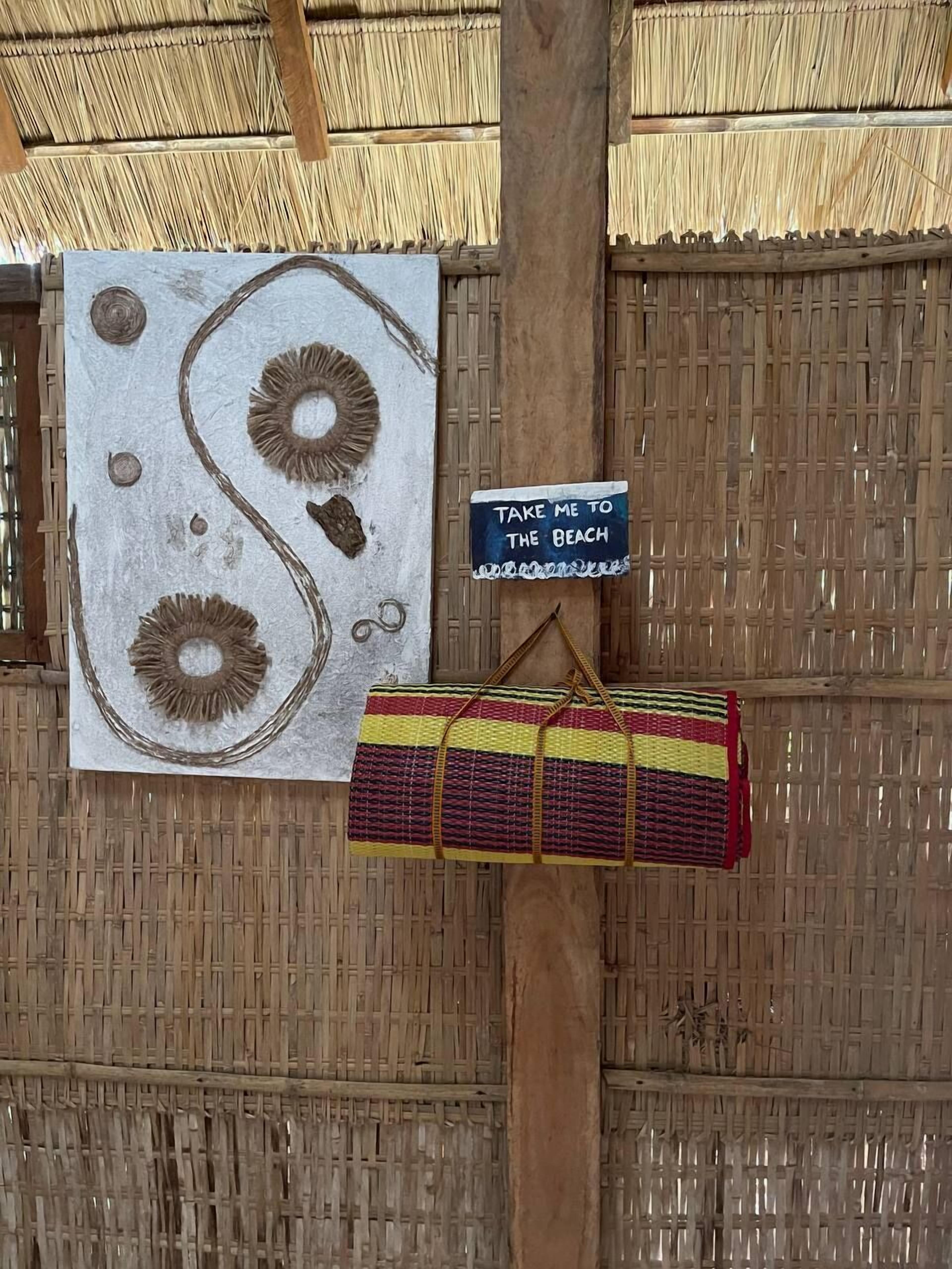 A room with a thatched roof, a wall-mounted sign saying "TAKE ME TO THE BEACH", and a woven bag hanging from a wooden post.