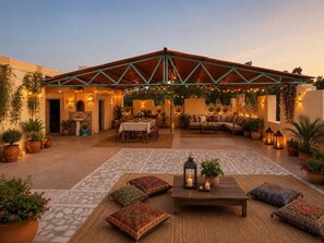 An outdoor patio area with a covered seating area, potted plants, and a table with cushions. - Hotel Robin Jaisalmer (Jaisalmer)