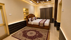 A bedroom with a bed, a nightstand, a wall-mounted shelf with decorative items, a patterned rug, and a ceiling with recessed lighting. - Hotel Robin Jaisalmer (Jaisalmer)