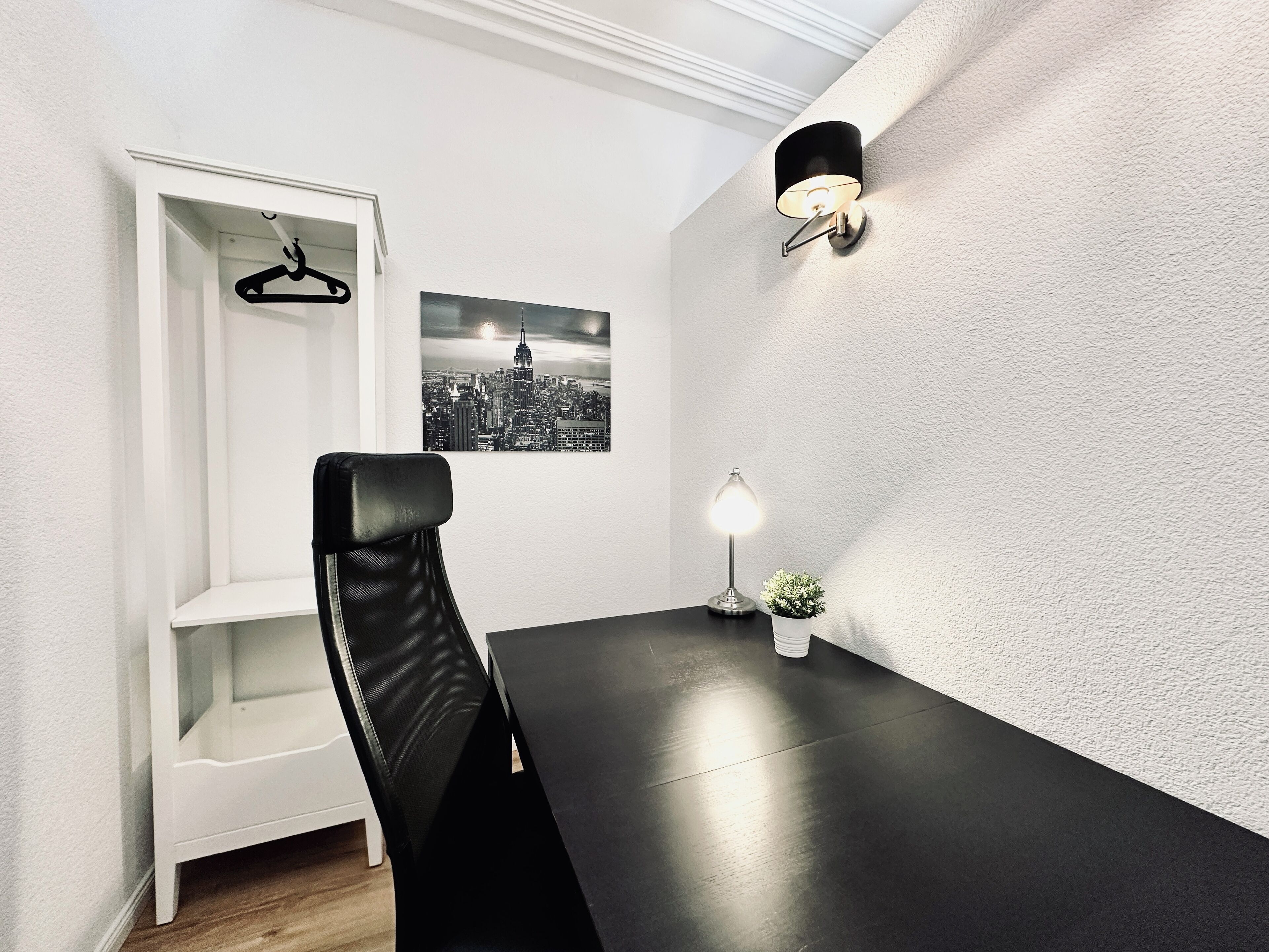 A modern dining area with a black table, a white chair, a wall-mounted lamp, a small potted plant, and a cityscape picture on the wall.