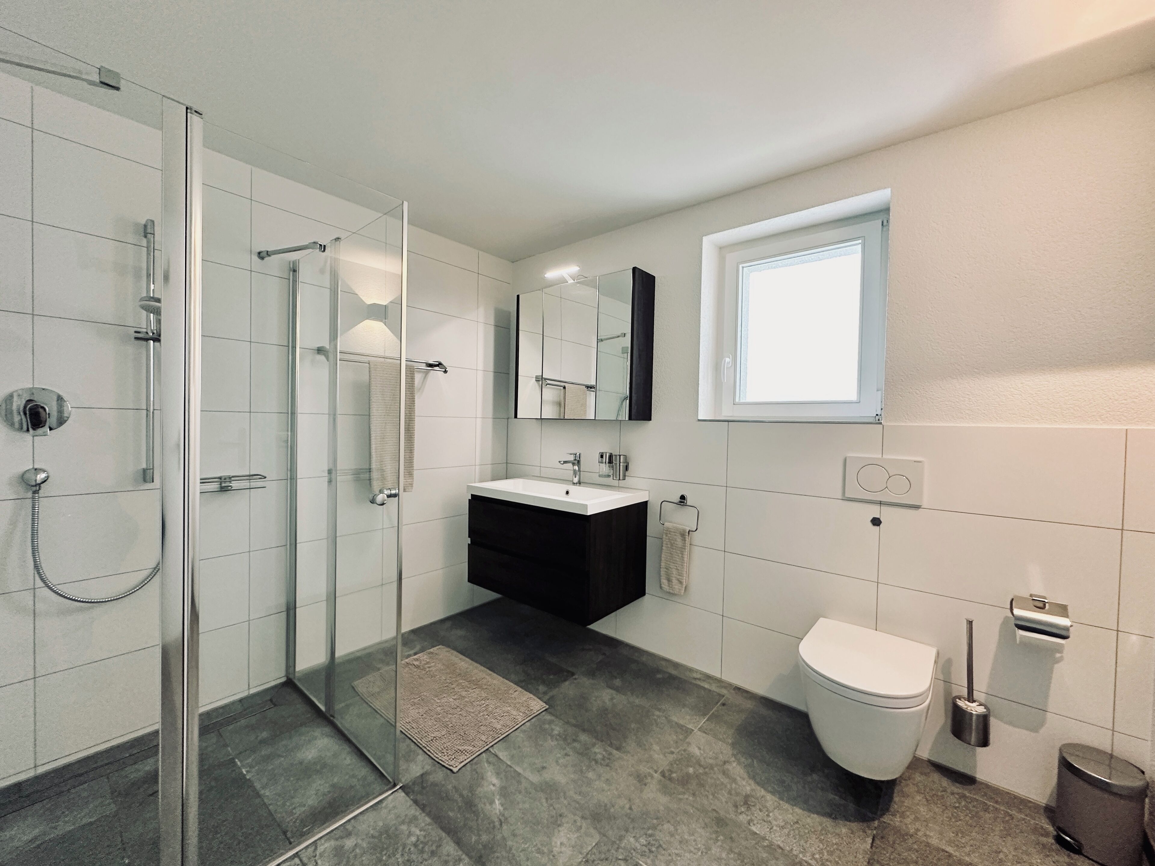 A modern bathroom with a glass-enclosed shower, a white sink, a dark vanity, a toilet, and a window.