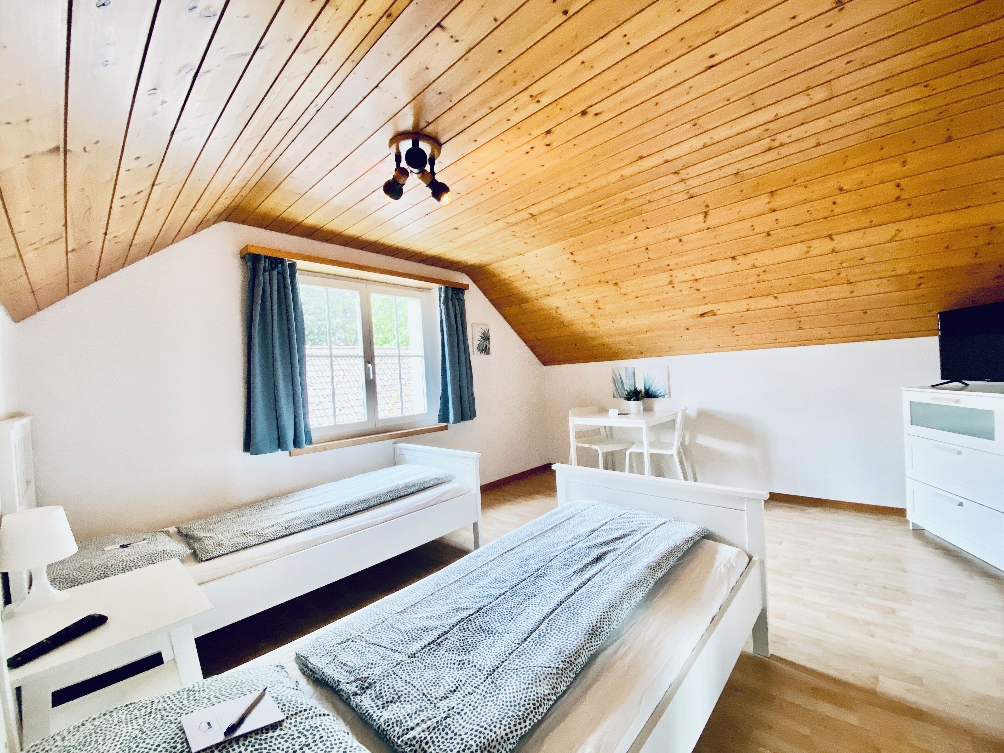 Um quarto de sótão aconchegante com teto de madeira inclinado, uma cama, uma escrivaninha e uma janela com cortinas.