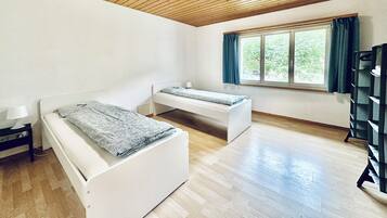 A room with two beds, a window with curtains, a wooden ceiling, and a bookshelf.