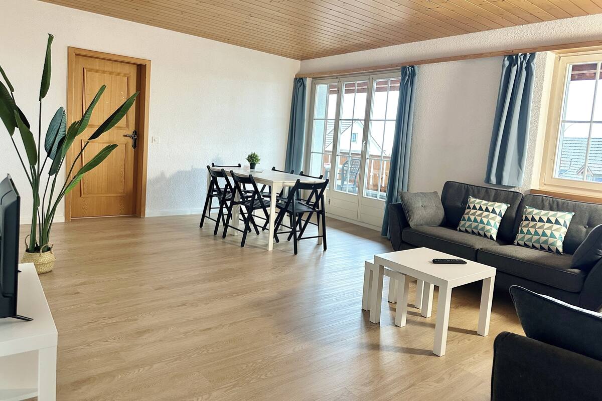 Ein geräumiger Wohnbereich mit Holzbalkendecke, einem Esstisch mit Stühlen, einer Couch mit Kissen und einem Fernseher auf einem Schrank.