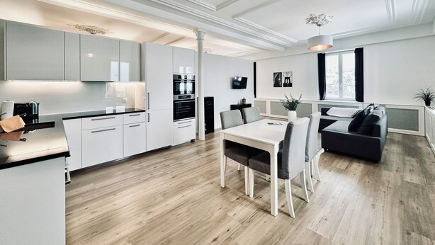 Een moderne keuken met witte kasten, een eetgedeelte met een witte tafel en stoelen, en een zithoek met een zwarte bank en een raam.