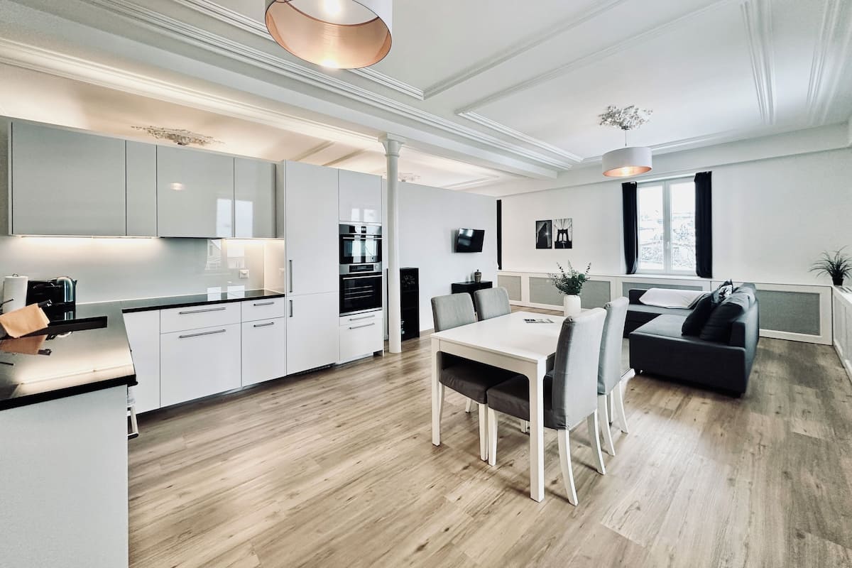 Eine moderne Küche mit weißen Schränken, ein Essbereich mit weißem Tisch und Stühlen sowie ein Wohnbereich mit einer schwarzen Couch und einem Fenster.