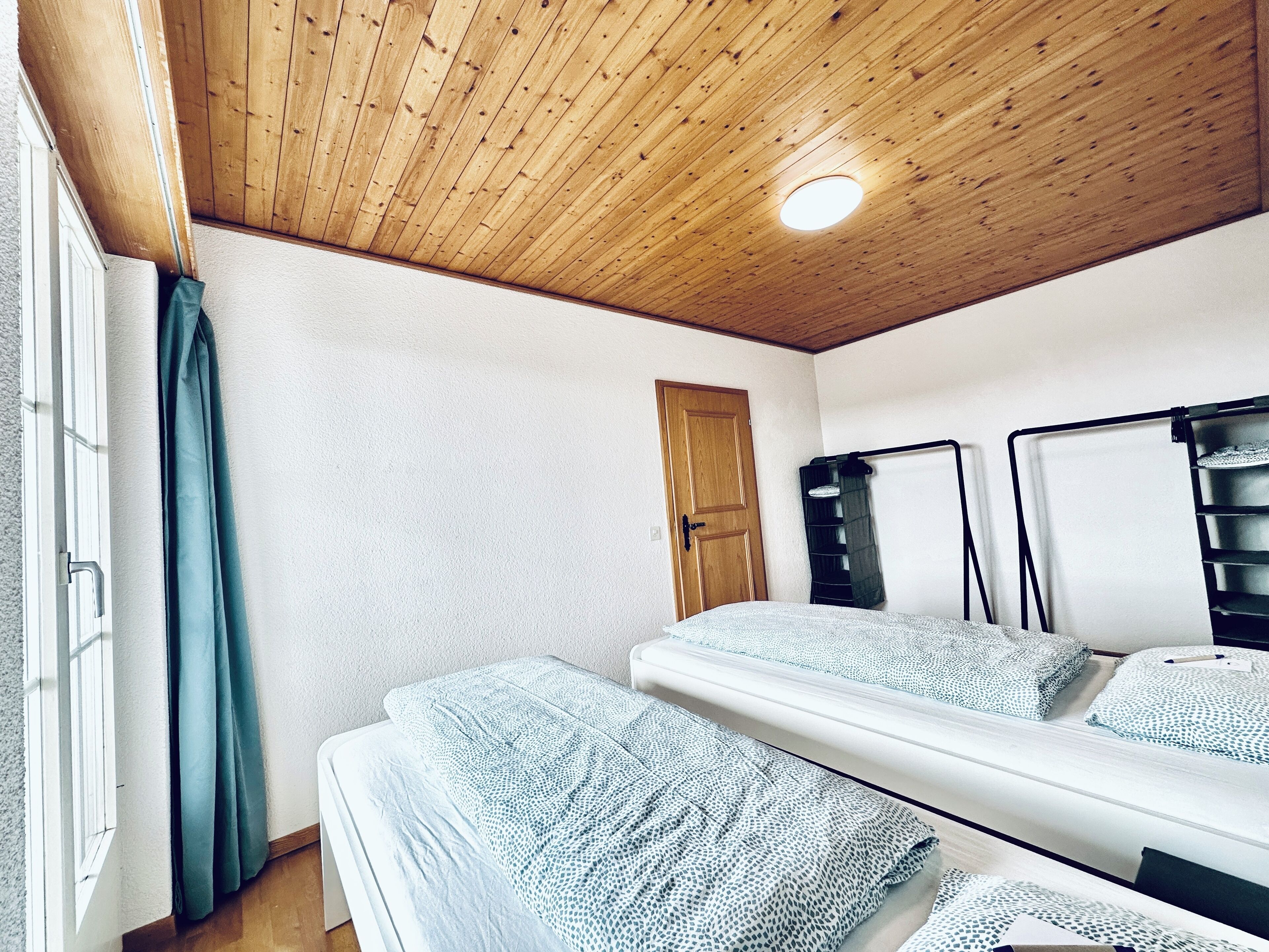 Une pièce avec un plafond en bois, une porte, une fenêtre avec des rideaux et un lit recouvert d’une literie à motifs.