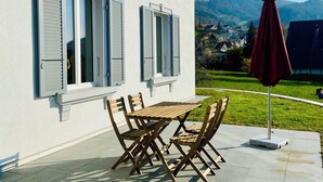 Een wit gebouw met luiken, een houten tafel en stoelen, en een rode parasol op een terras.