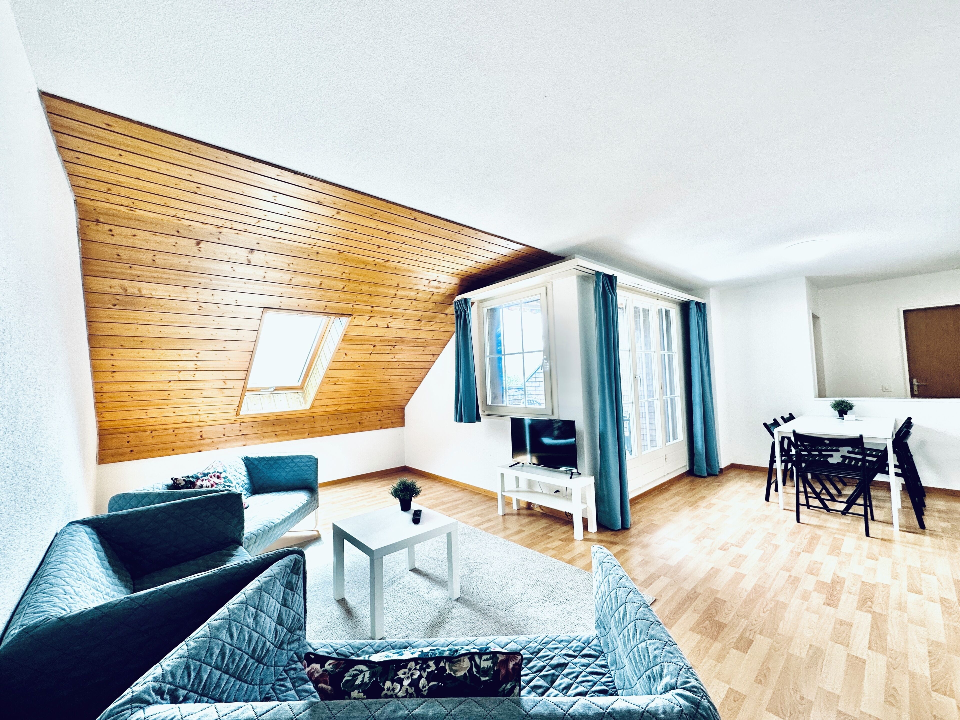 Una sala de estar moderna con techo inclinado de madera, un sofá, una mesa de centro, un televisor y una zona de comedor con sillas y una mesa.