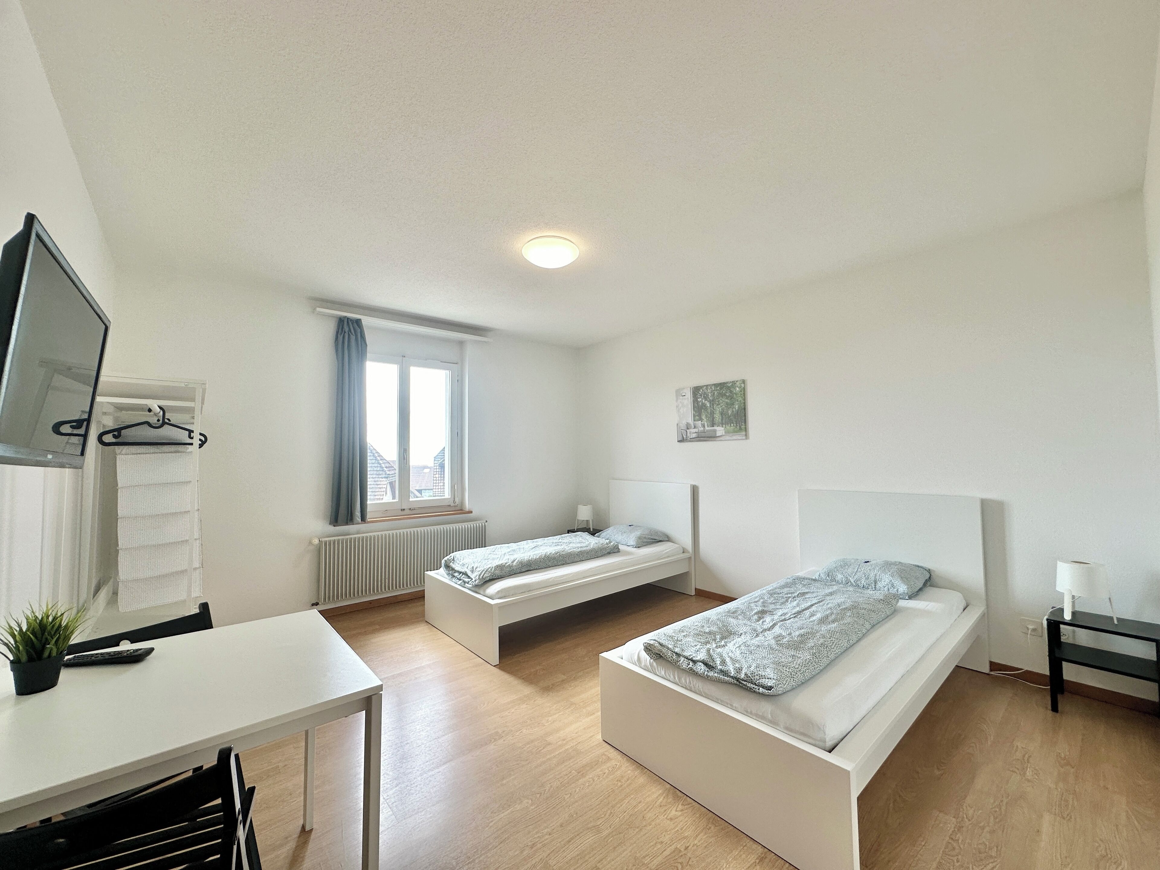 A modern bedroom with two single beds, a desk, a radiator, and a window with curtains.