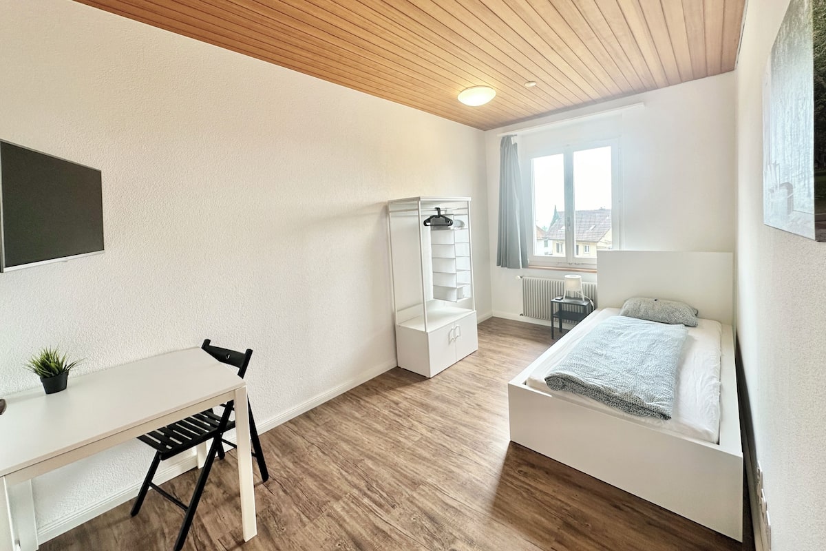 Ein Schlafzimmer mit Bett, Schreibtisch, Stuhl, Kleiderschrank und einem Fenster mit Blick auf Gebäude.
