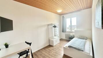 A bedroom with a bed, a desk, a chair, a wardrobe, and a window with a view of buildings.