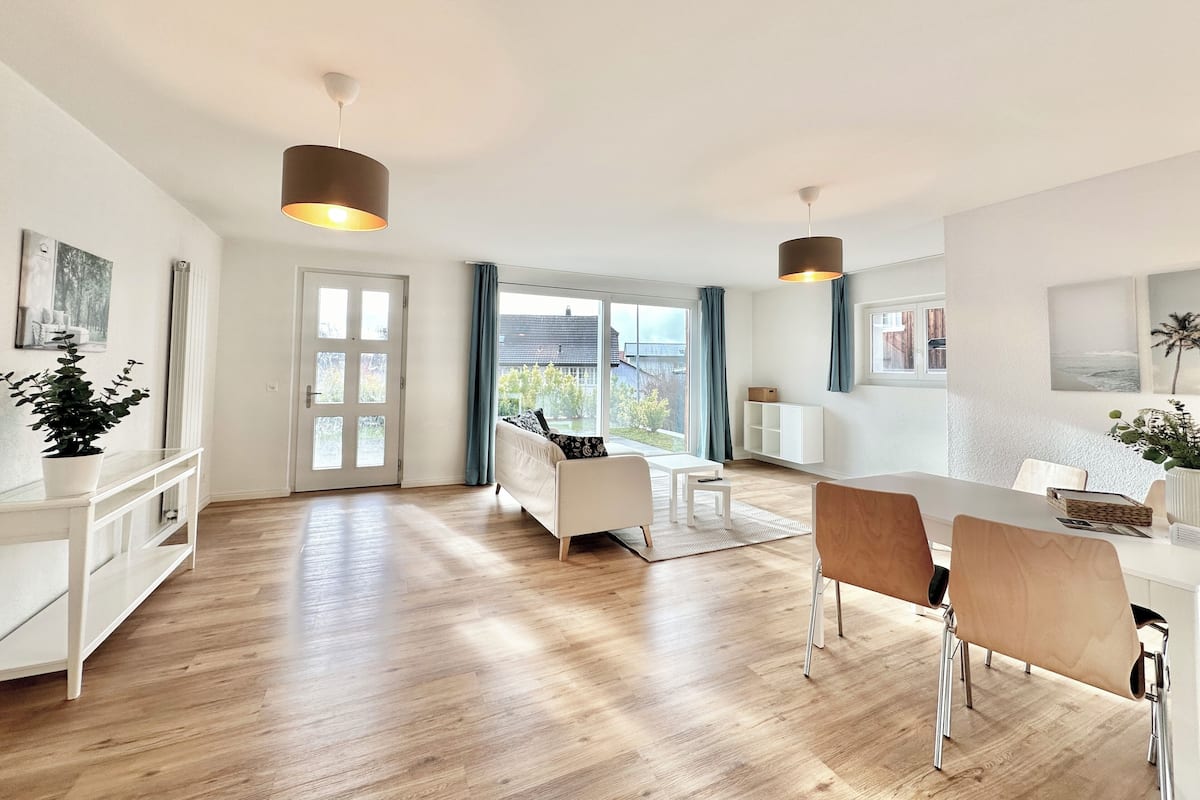 Ein großzügiger Wohnbereich mit Esstisch, Stühlen und einer Couch. Ein großes Fenster bietet Blick ins Freie.