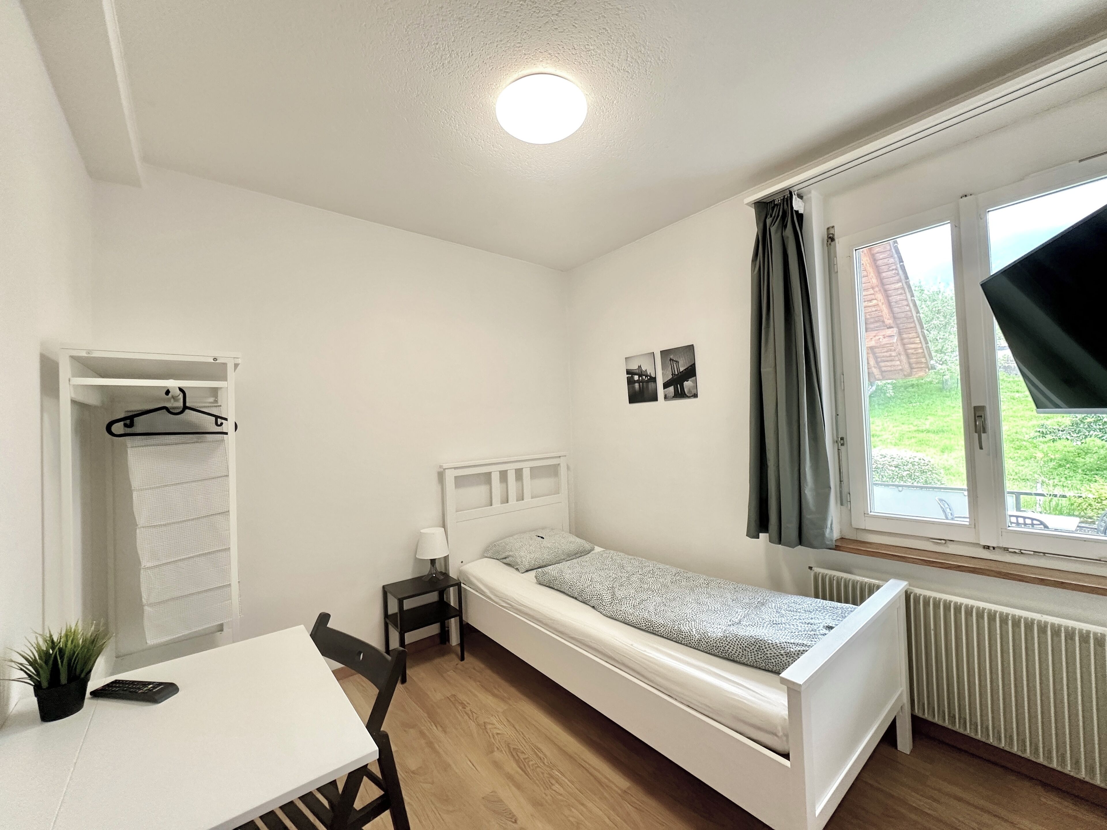Une petite pièce bien rangée, avec un lit simple, un bureau, une chaise, une petite table, une table de chevet, une plante en pot et une fenêtre avec des rideaux.